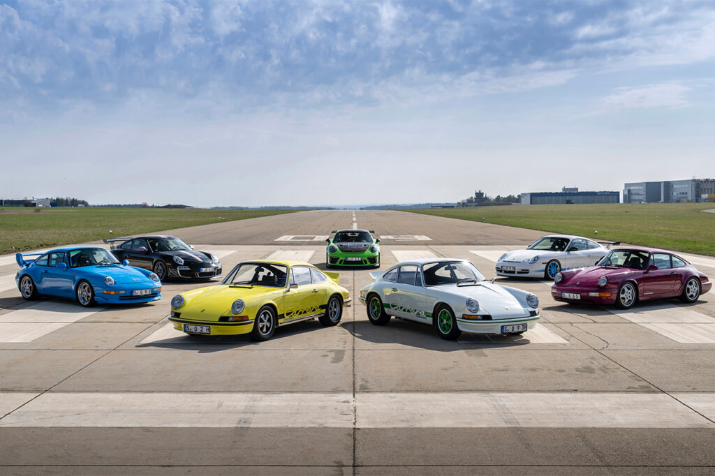 Generations of 911 RS cars parked next to one another on a strip of road with grassy fields to the left and right and blue sky with clouds in the background