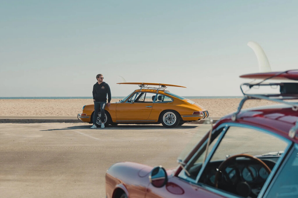 Guy leaning on the hood of a yellow Porsche 911 with a surfboard strapped on top and a red porsche staged in front of the yellow one