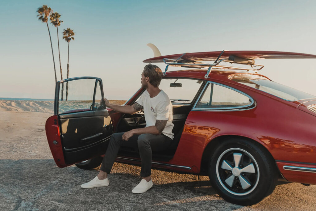 Man sitting in side of Red porsche with a surfboard on the roof with palm trees in the background