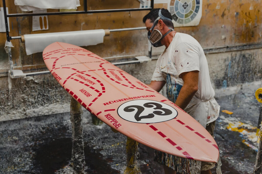 "Pink Pig" Porsche livery surfboard that a man is standing next to inside of a shop