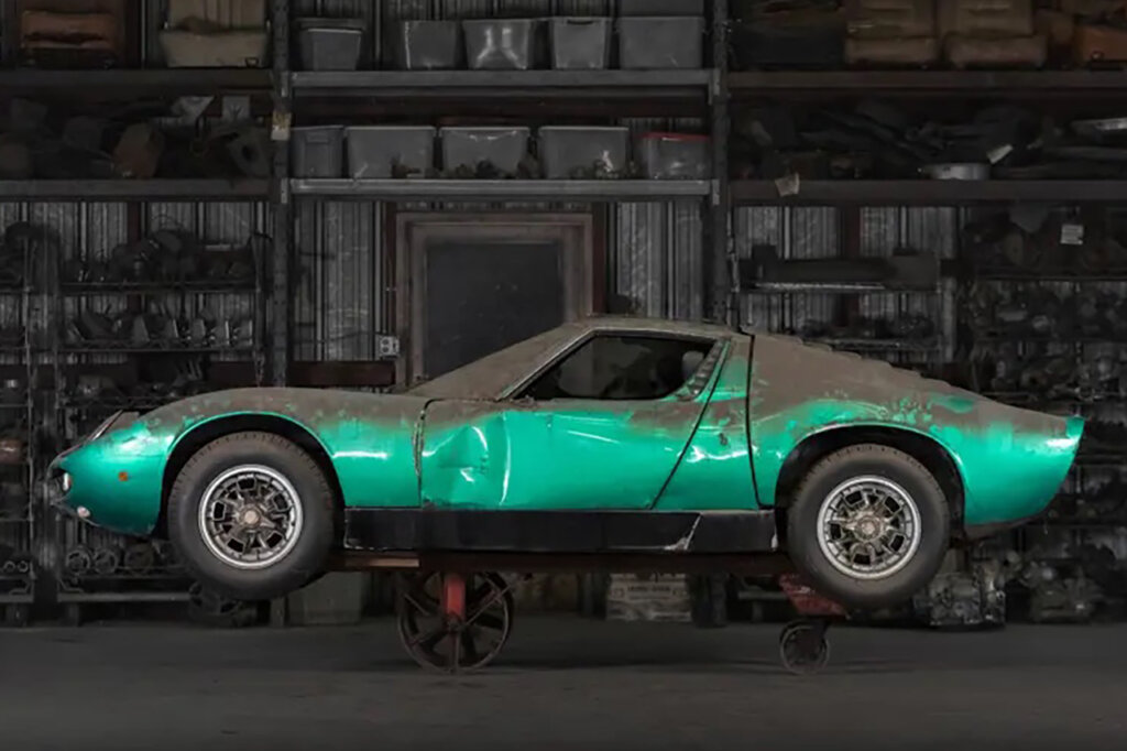 Dirty dust covered green Lamborghini Miura P400 propped on a metal cart with wheels in a garage with shelves in the background