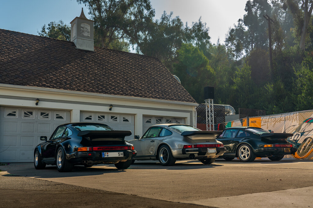 Three 911's 2 black, 1 silver parked in a driveway of a house with a fence covered in a banner behind one of the cars