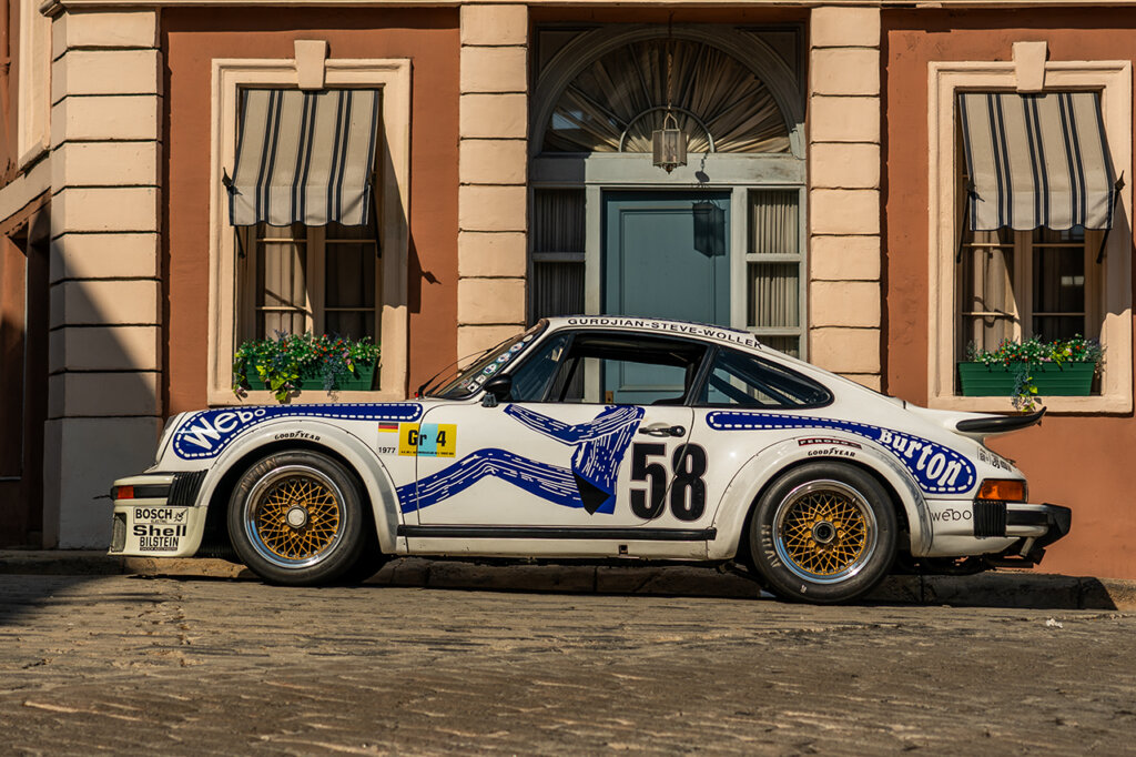 White and blue 911 934 parked on a stone road in front of a brown building with striped window covers