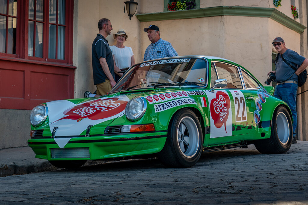 Green and white Porsche race car parked next to a sidewalk with spectators standing near the car