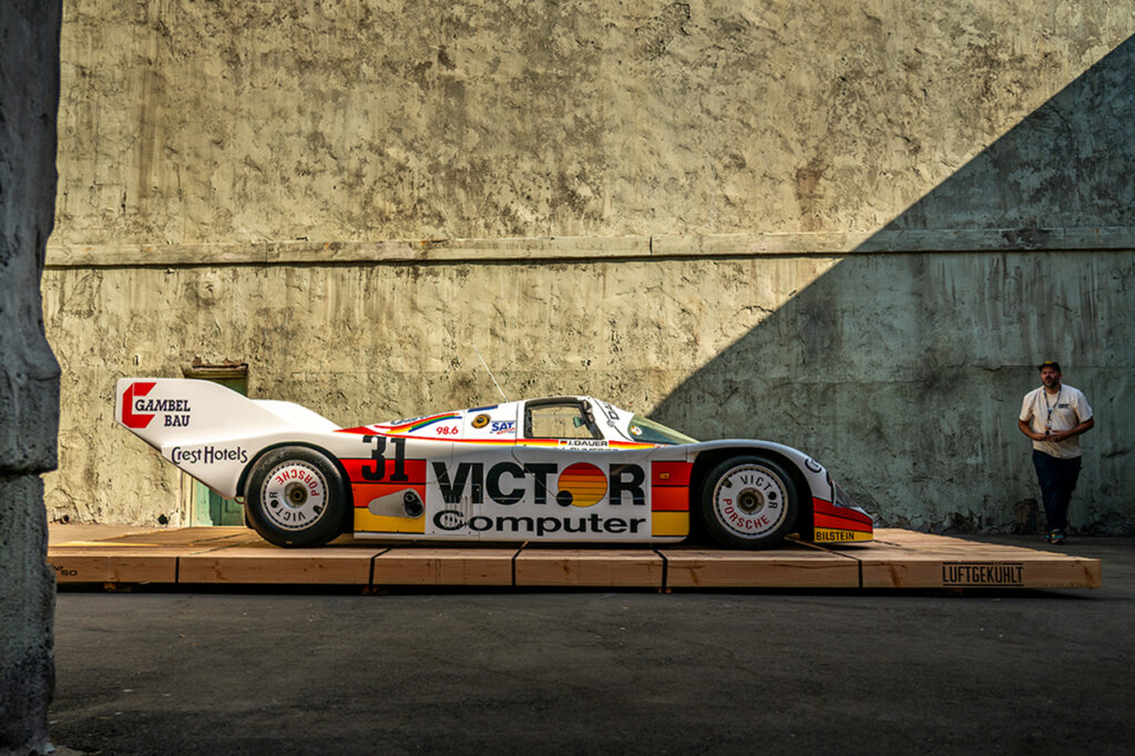 Orange and white Porsche 962 with race car livery that says Victor Computers on the side postitioned on a wooden platform along a textured wall with a shadow of a line crossing through