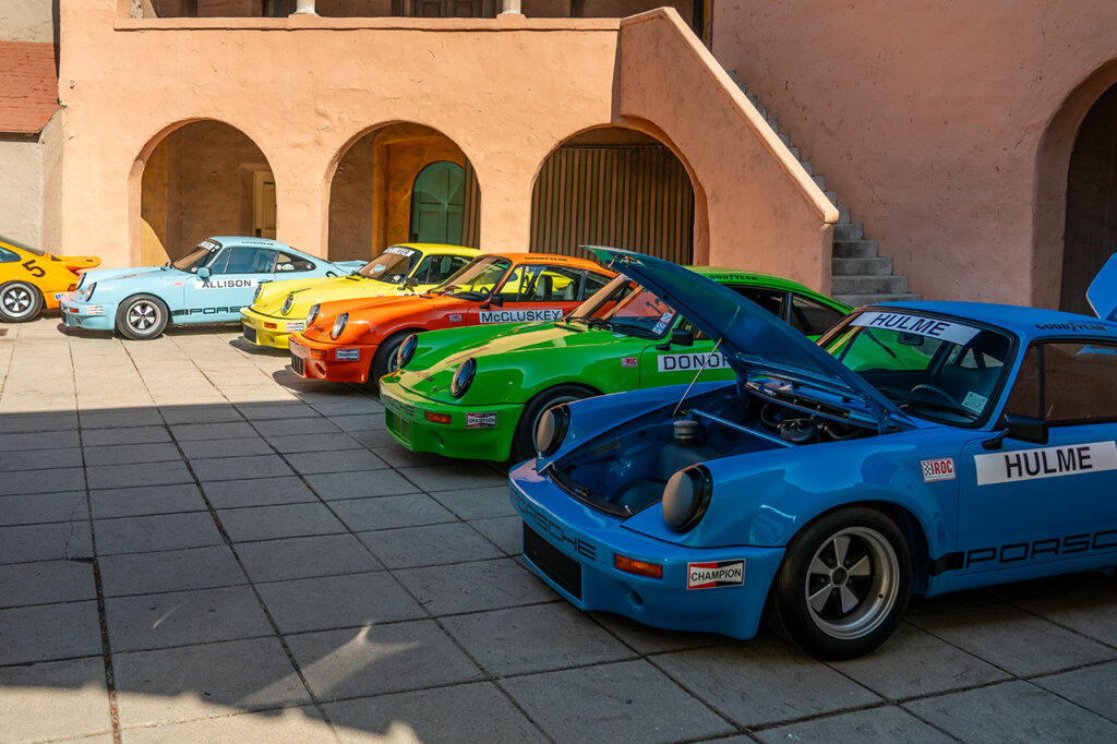 Blue, green, orange and yellow Porsche 911's parked next to one another in a courtyard surrounded by an orange building