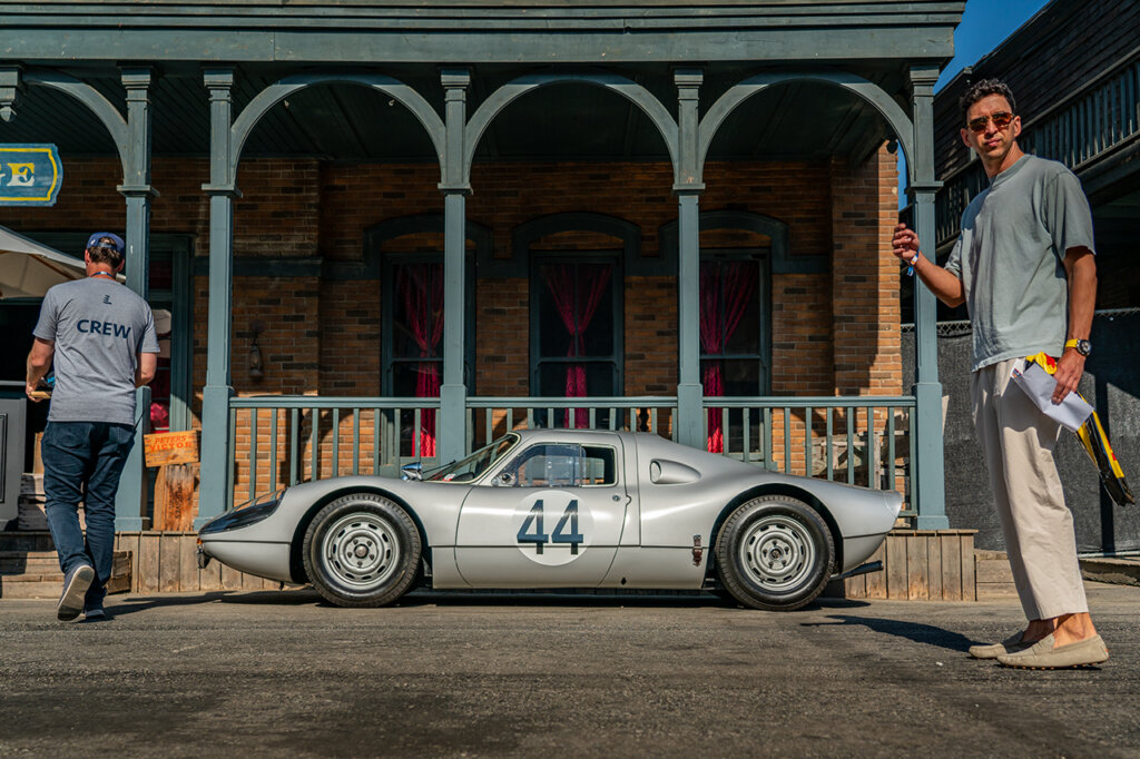 Silver grey Porsche 904/6 with the number 44 in a white circle on the door parked on the side with two men standing on either side of the car in front of a green porch