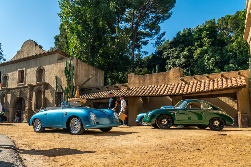 Two Porsche 356's staged on a dirt path one blue and one dark green with many people walking around both of these cars