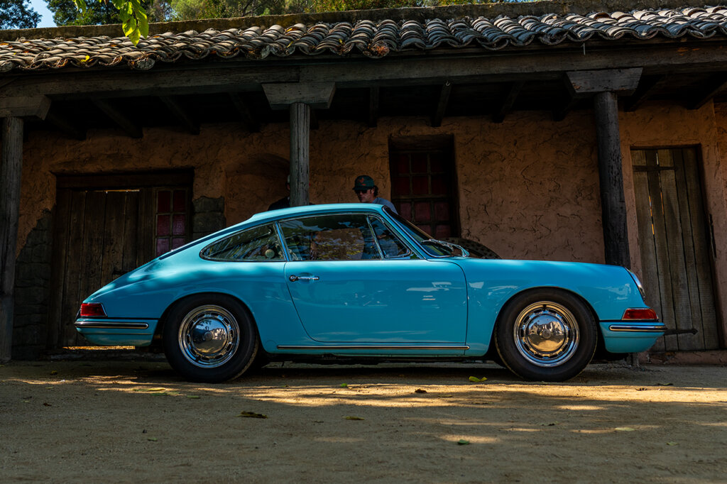 Baby blue 912 sitting on a gravel road with a wooden building behind it and two people standing next to the driver door