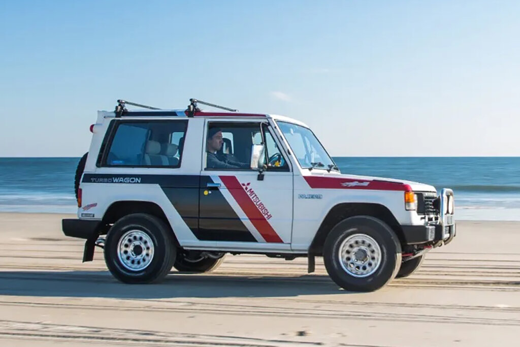 White, red and black Pajero suv driving on the sand with the ocean in the background