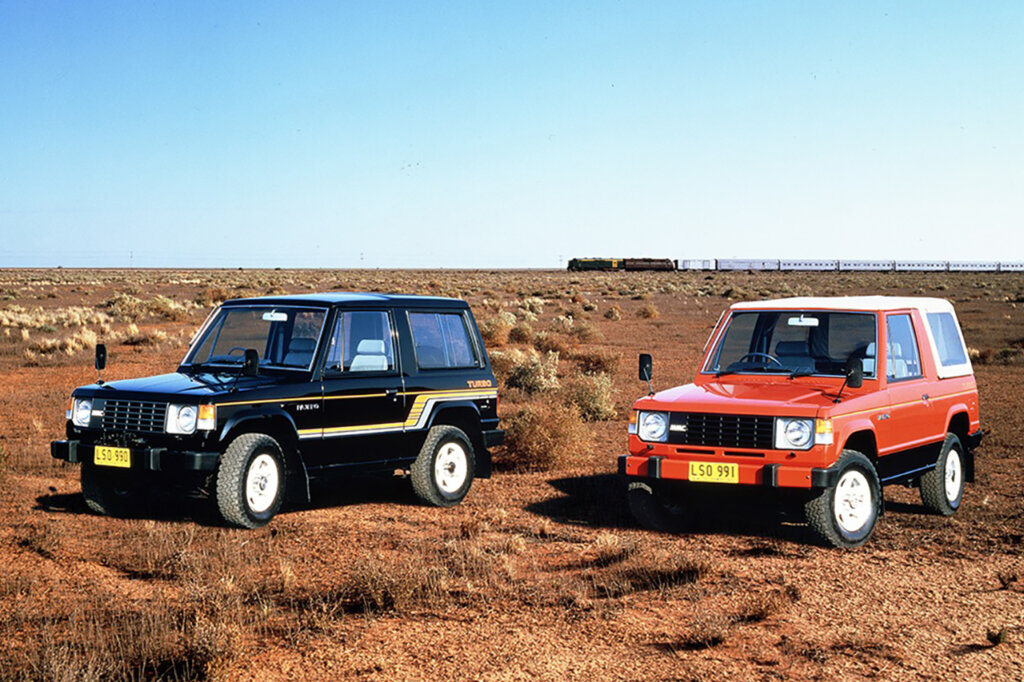 Two 1st gen Mitsubishi Pajeros's one black and one red next to each other on a dry red grassy field