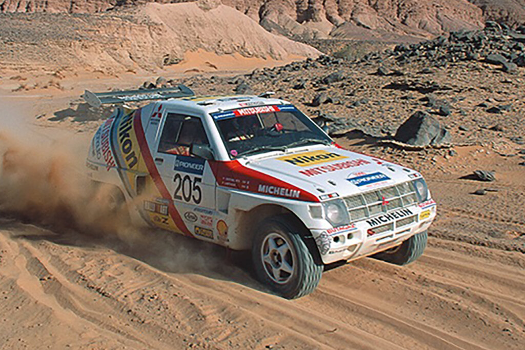White red and black mitsubishi pajero driving through sand with hills and trees in the background