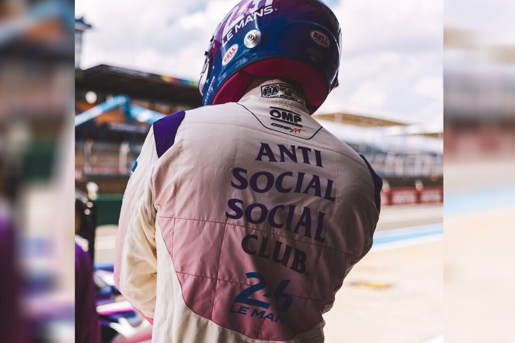 Race car driver wearing a cream and purple gradient racing suit with the brand Anti Anti Social Club branded under the OMP logo and 24hr Le Mans logo