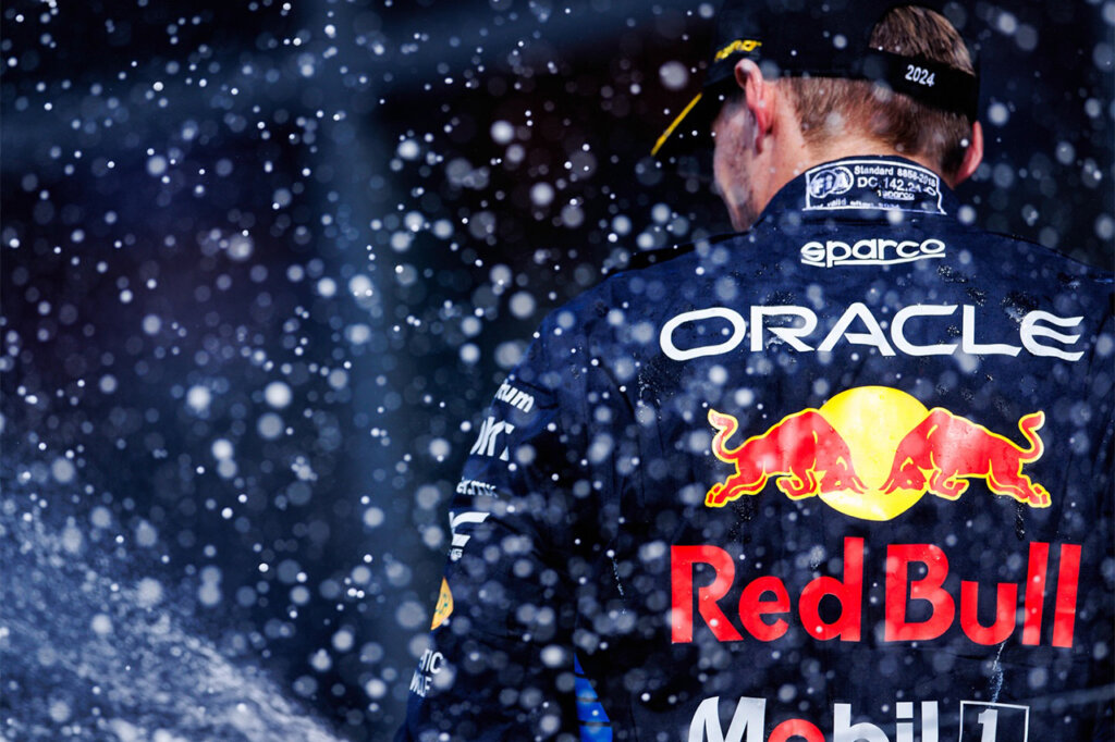 Man wearing a hat with a blue racing suit with the brands Sparco, Oracle, Red Bull and Mobil 1 adorning the back of the suit with water droplets covering the surrounding area