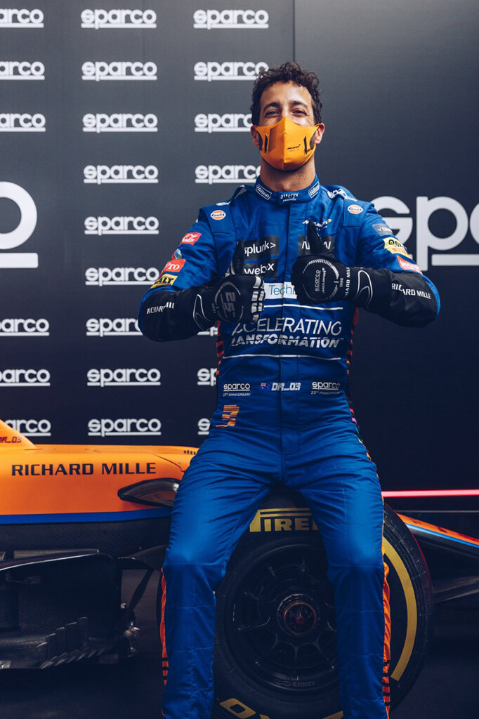 Sparco F1 driver Daniel Ricciardo posing with thumbs up sitting on an orange F1 car and wearing an orange mouth covering