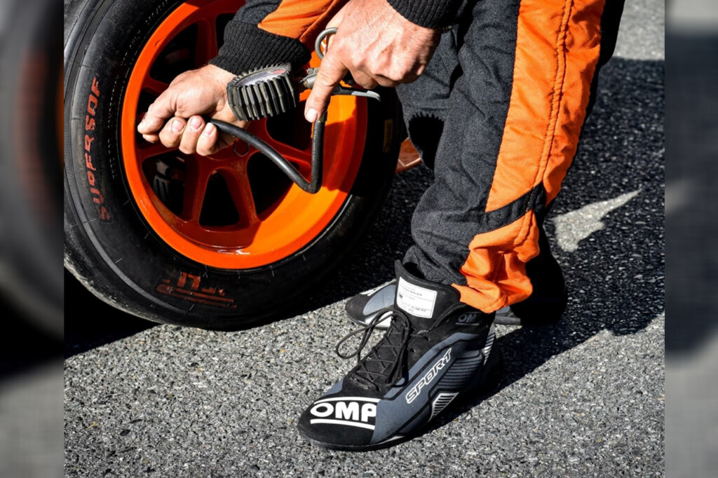 Man holding a tire gauge wearing black leather OMP safety shoes checking tire pressure of a wheel with an orange rim