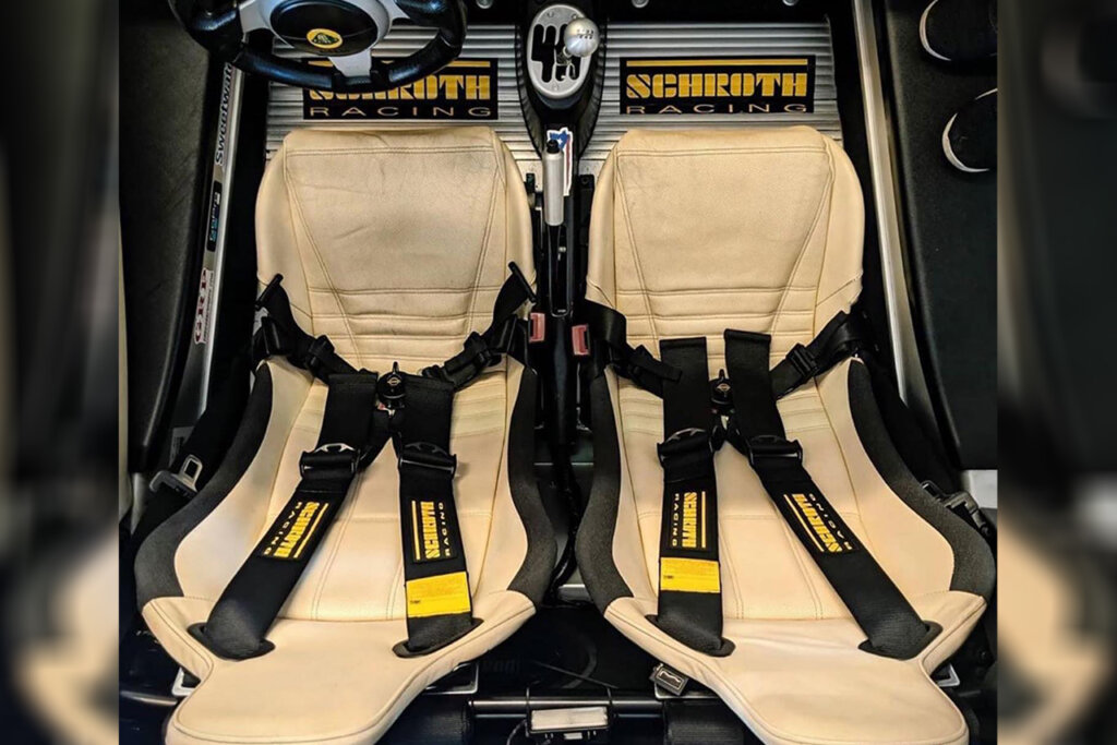 Overhead view of a car's interior with beige seats, black Schroth harness safety belts and with black and yellow Schroth branding on floorboard