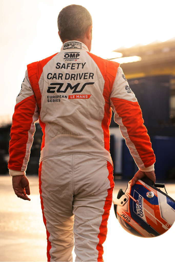 man with an orange and white racing suit holding an orange and black helmet walking away with the sun setting in the background