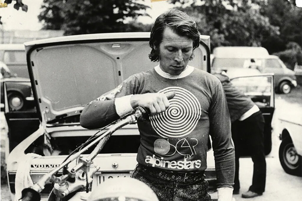 Black and white photo of a man wearing an Alpinestars long sleeve tshirt as he stands next to a motorcycle