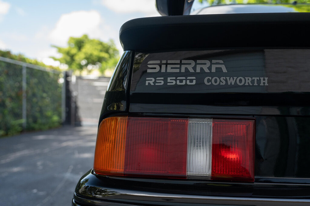 Black car with Sierra RS500 Cosworth badge above red and orange taillights with green trees and blue sky in background