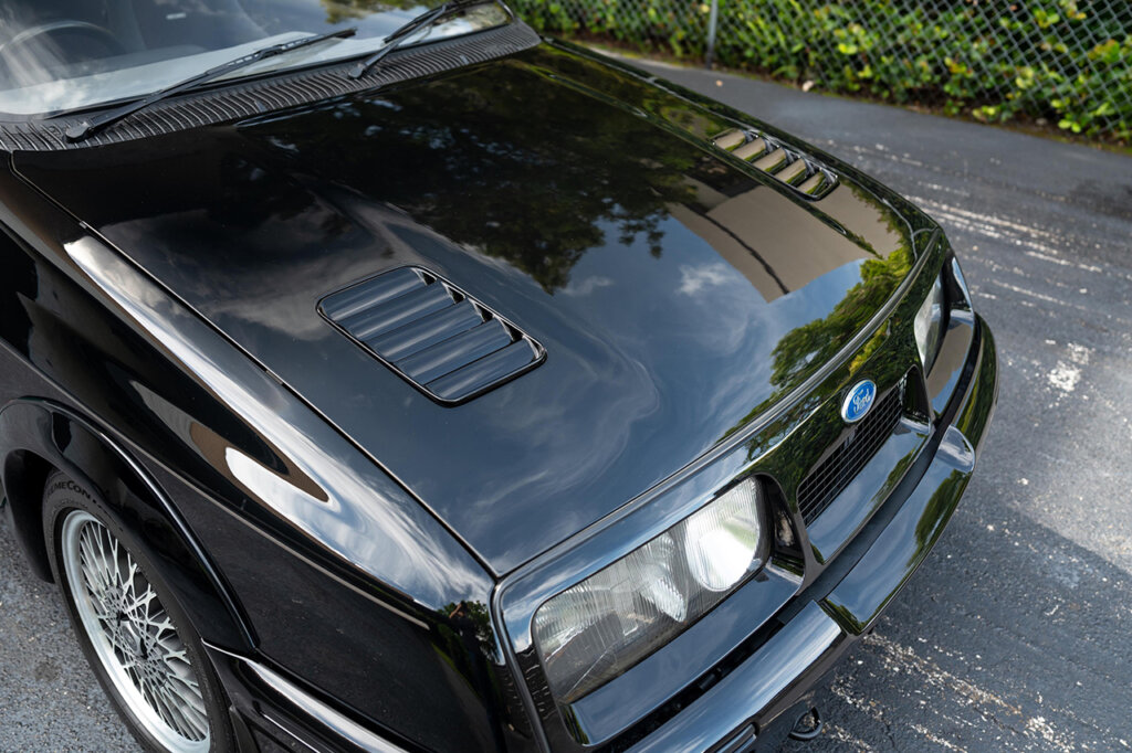 Black Ford Sierra RS500 with black hood vents with clear headlights next to a blue oval Ford badge