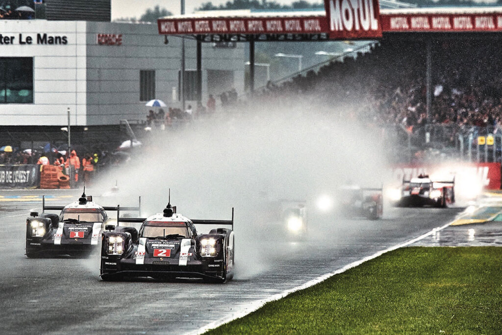 Hypercar race cars driving on a wet race track leaving a spray of water behind it with cars behind one another.