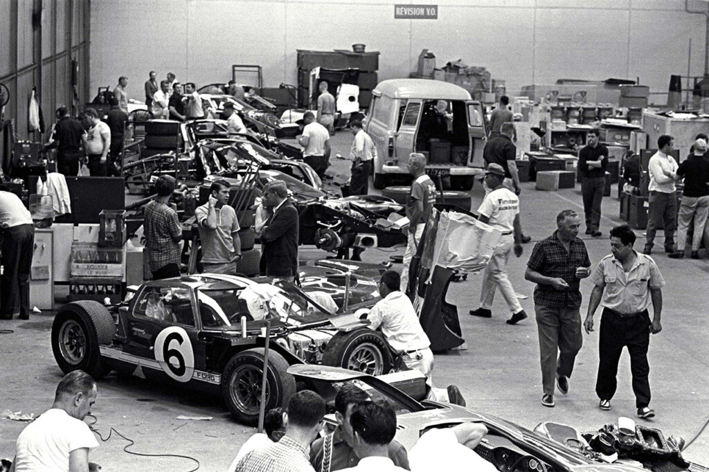 Black and white photo of Ford race cars in a garage with many people rushing between cars working on them