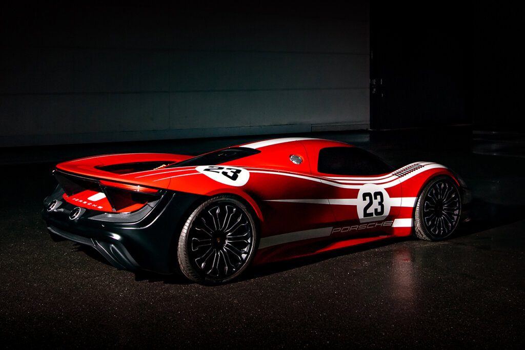 Red and white Porsche 917 Living Legend parked in a dark room with the car under a spotlight with white brick wall behind it.