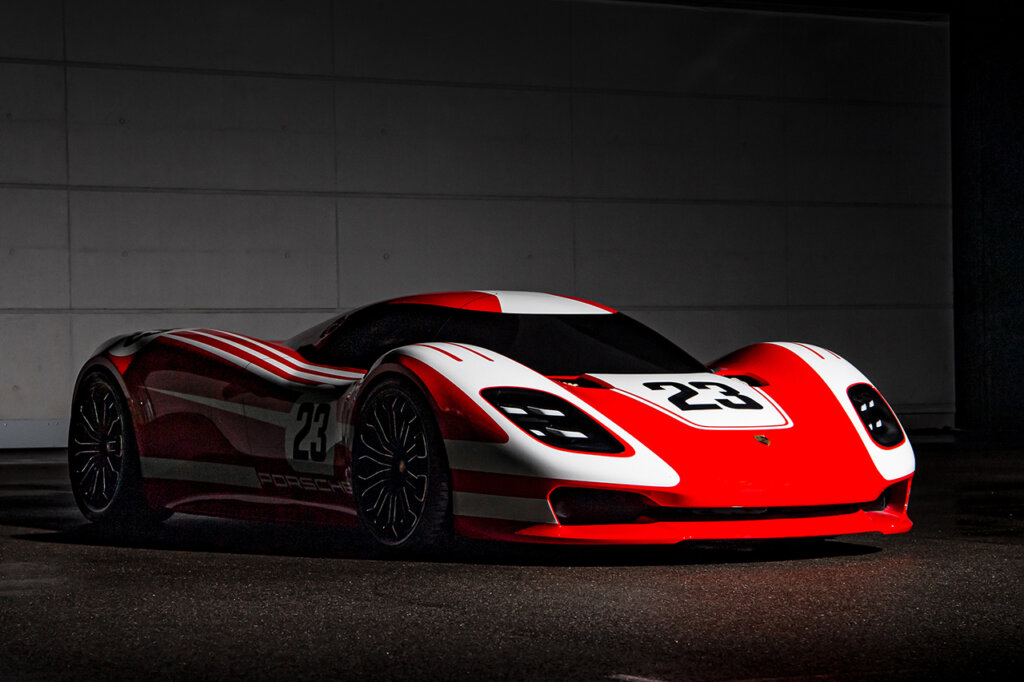 Red and white Porsche 917 Living Legend parked in a dark room with the car under a spotlight with white brick wall behind it.