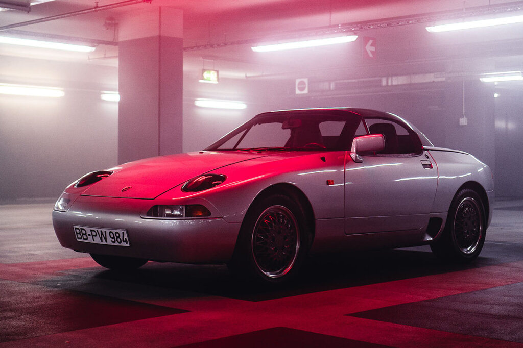 Silver Porsche 984 concept car in a garage with rows of lights above and a red spotlight lightly covering the car in a glow