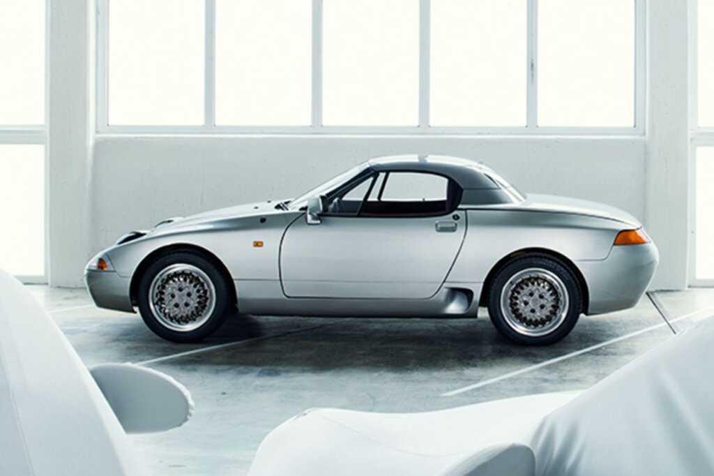 Side shot of a Porsche 984 concept car in a white room with large windows and two cars in the foreground under car covers.
