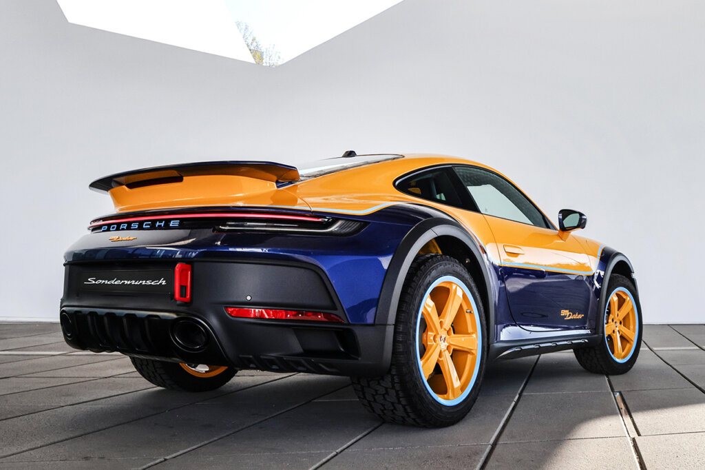 Yellow and blue Porsche 911 Dakar sitting in a room with white walls and concrete square flooring with a skylight.