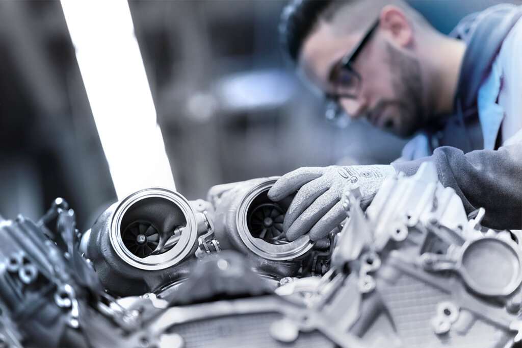 Man wearing black glasses adjusting two turbochargers on an engine inside of a factory with a light above him.
