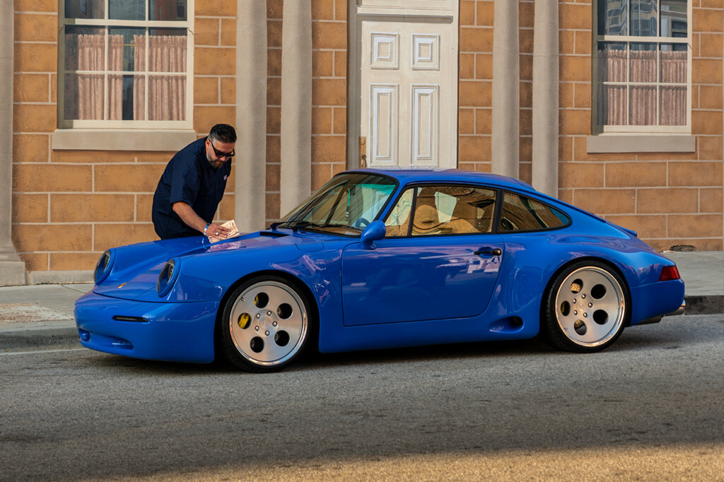 Blue modified Porsche 993 parked on the side of the road with a man wiping down the hood and large polished wheels.