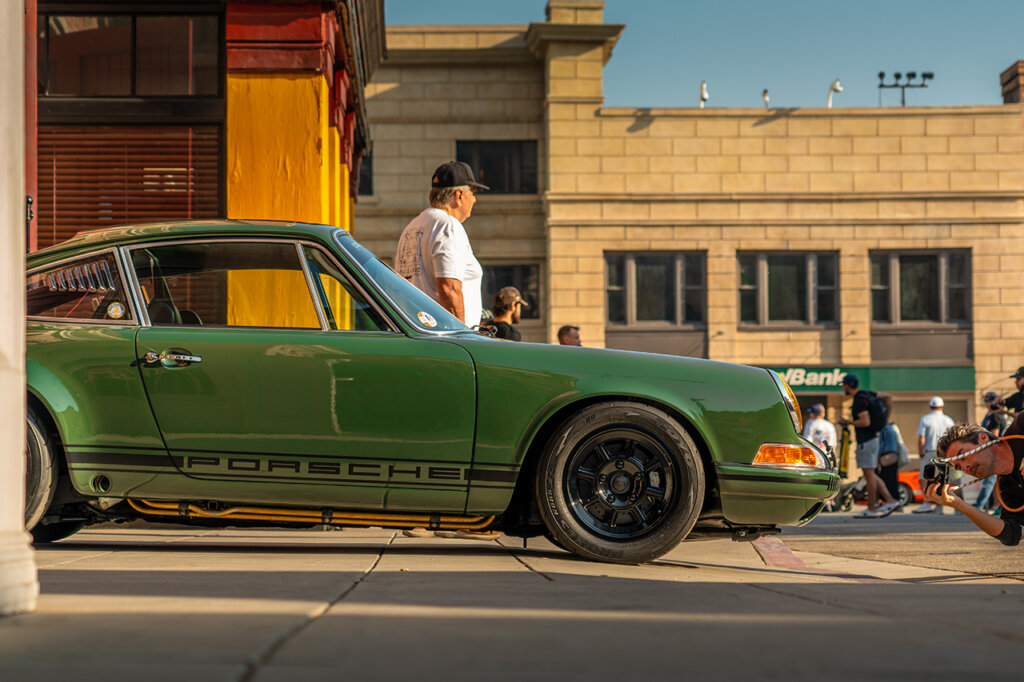 Green 911 parked extending through a sidewalk with people passing and taking photos of the car