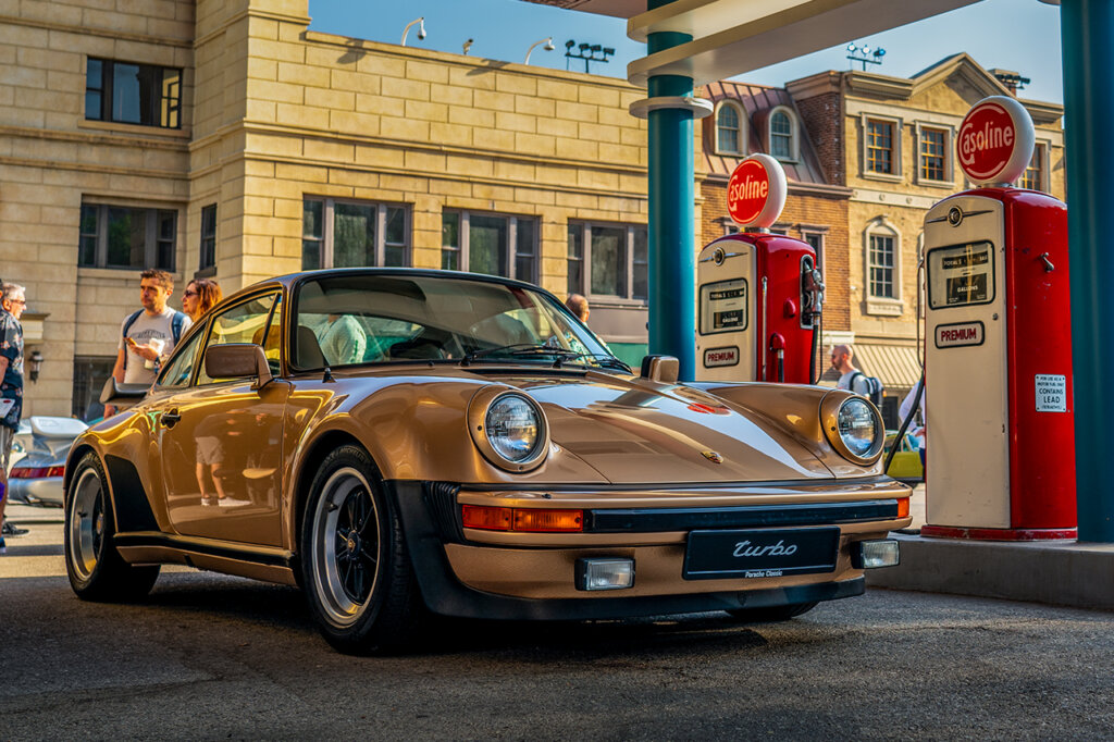 Bronze Porsche 930 Turbo parked in a staged gas station with two red gas pumps on the driver side