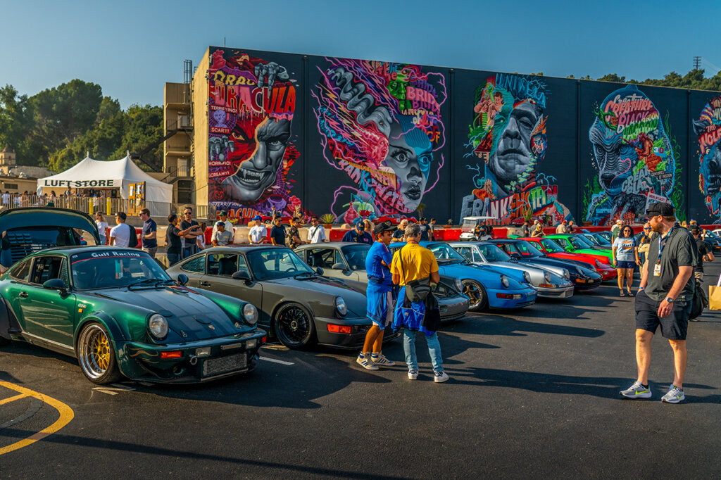 Several Porsche 911's green, blue, brown, grey, red staged in a line in a parking lot surrounded by many people walking and in front of a large colorful mural with characters on them