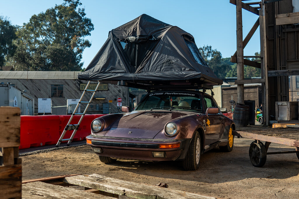 Overland Purple 911 with a camping tent on the top with wooden structures near the car