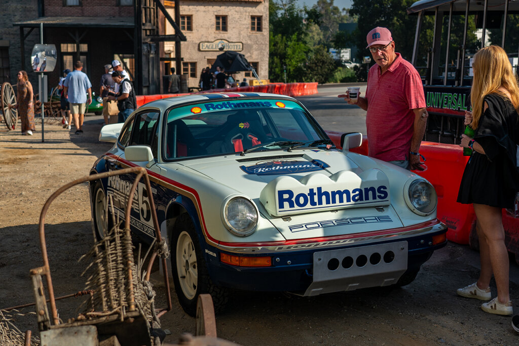Rothmans off-road 911 race car parked with several people walking really close to the car with a brown building in the background