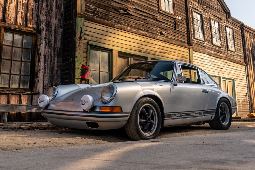 Silver 911 with two extra highlights on the hood with white covers parked in front of a wooden building