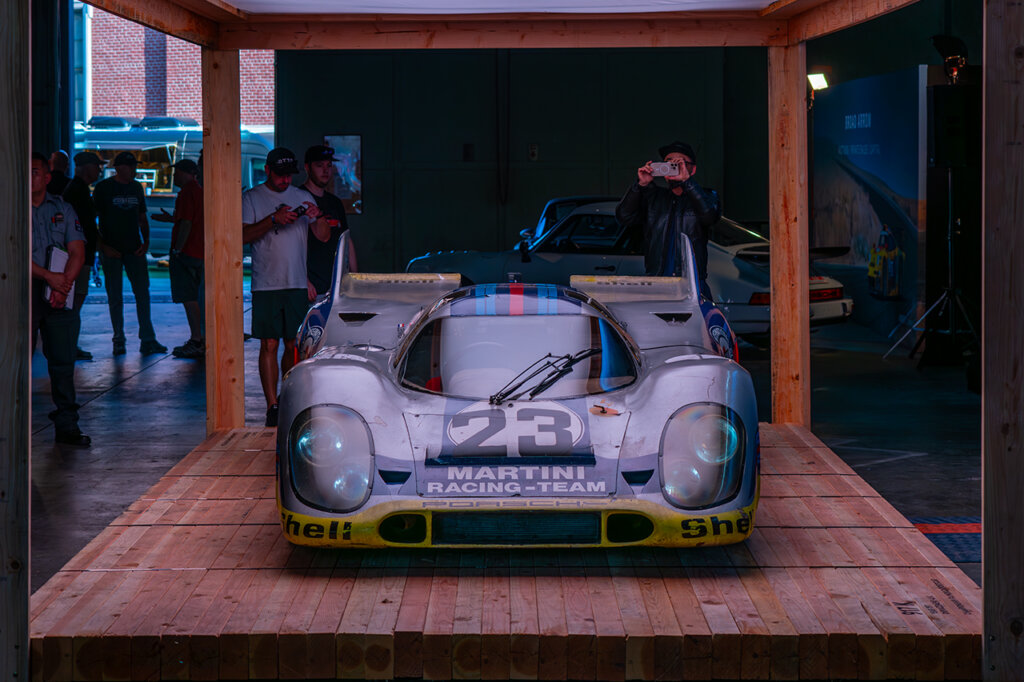 Silver Martini Porsche 917K race car parked on a wooden platform structure with many spectators around it and taking photos