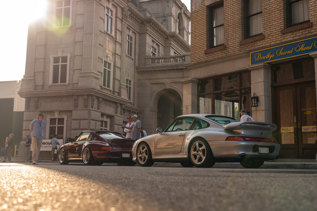 Two Porsche cars one silver and one purple parked on the street with the sun setting in the background going behind  tall stone buildings