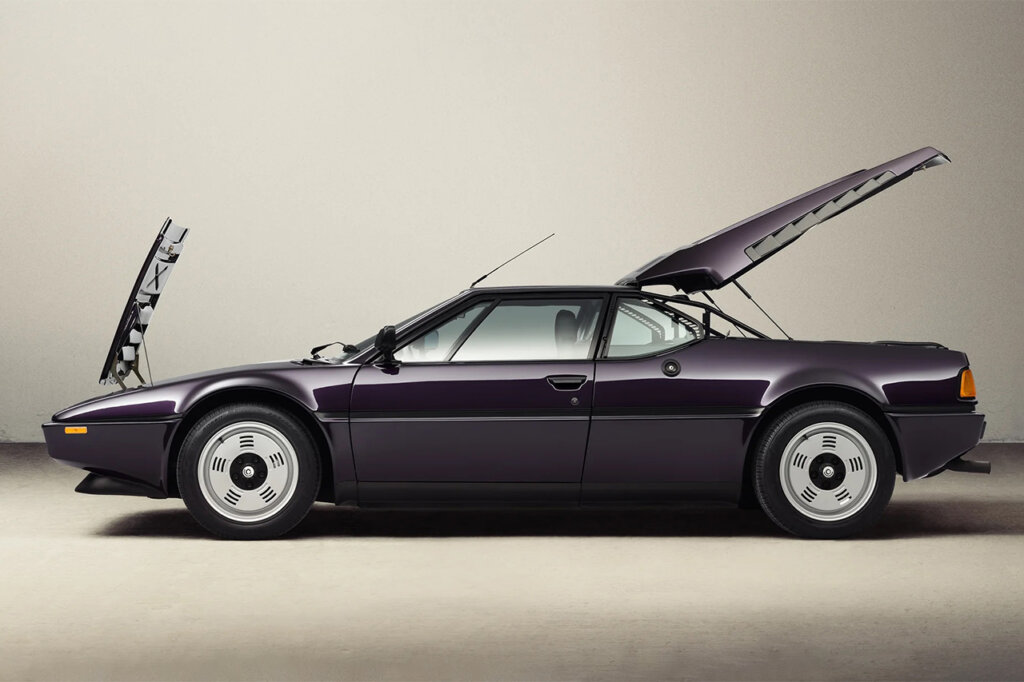 Techno Violet BMW M1 E26 by Ronnie Fieg & KITH shot sideways with hood and rear hatch propped open positioned in front of a beige wall and off white concrete wall.