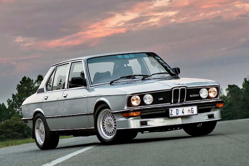 Silver BMW E28 M535i sedan with headlights on parked at an angle with sun setting behind clouds in the sky in the background.