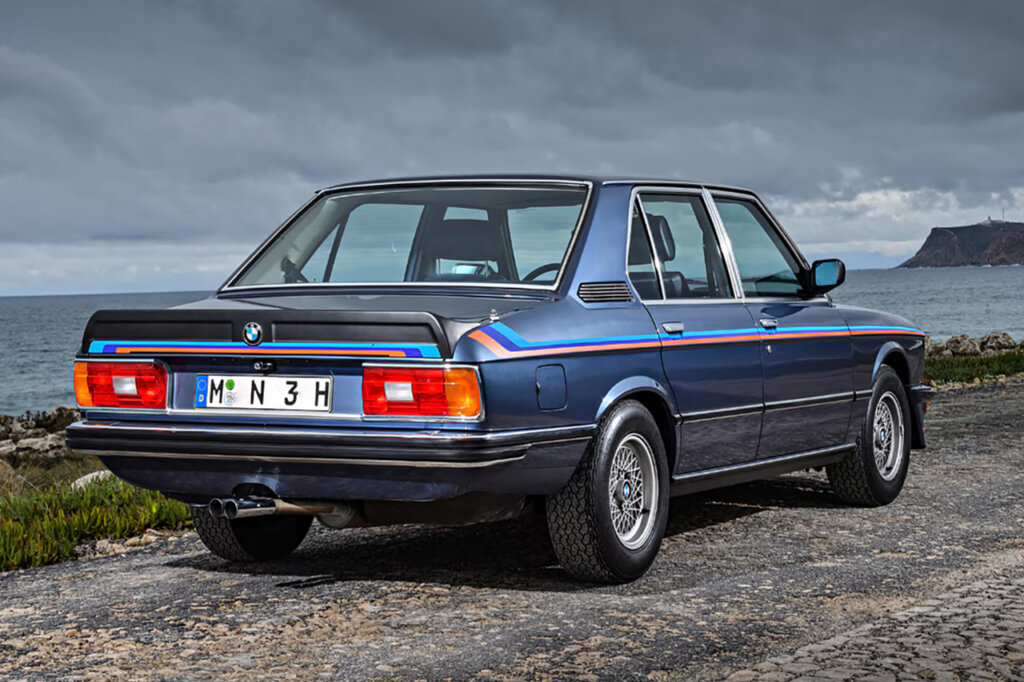 Blue BMW sedan with M division stripe going along the side of the car to the trunk with dark grey clouds in the background.