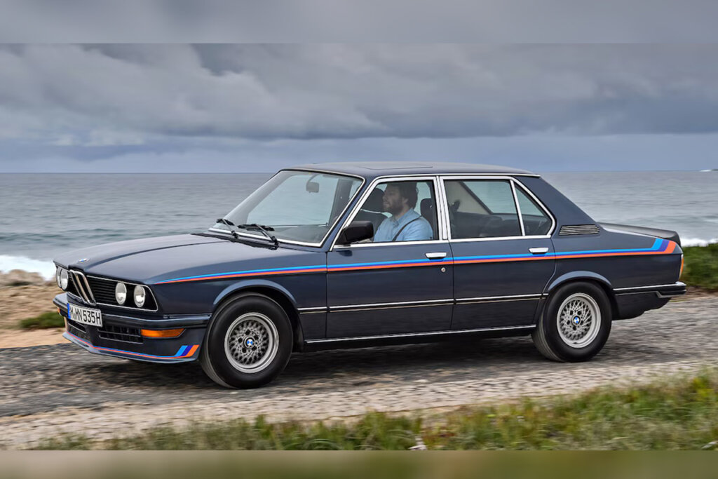 Blue E12 M535i sedan on a sandy road passing grass in the background and the ocean behind the car.