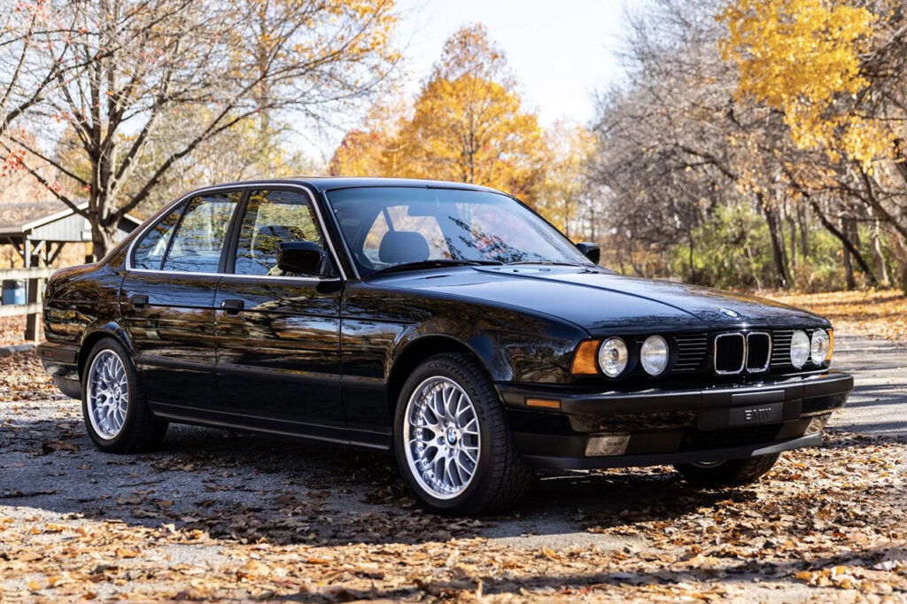 Black Saloon BMW M5 parked on a road with trees in the background and leaves on the ground.