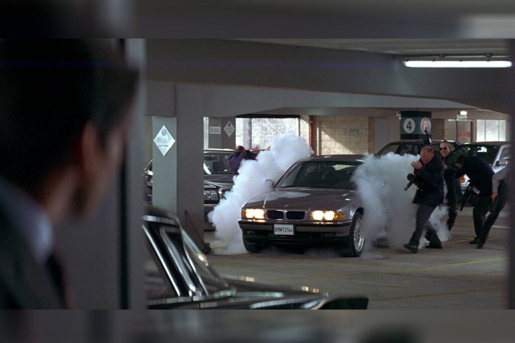 Man in a suit in the foreground with a grey BMW 750iL building tire smoke with several men next to it inside of a parking garage.