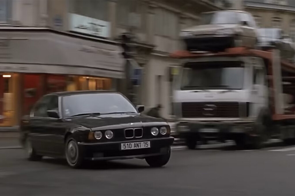 Black 1991 BMW M5 sedan overtaking a truck carrier on a road with buildings in the background.