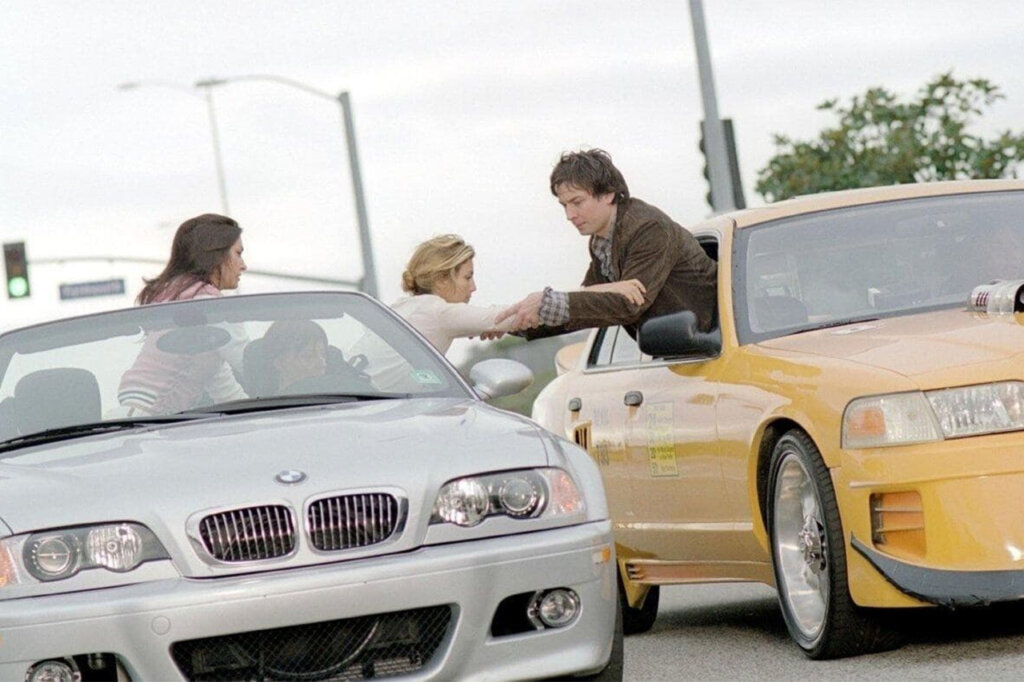 Silver BMW E46 Cabrio and modified yellow Ford Crown Victoria with people hanging out of the windows and reaching out to each other.
